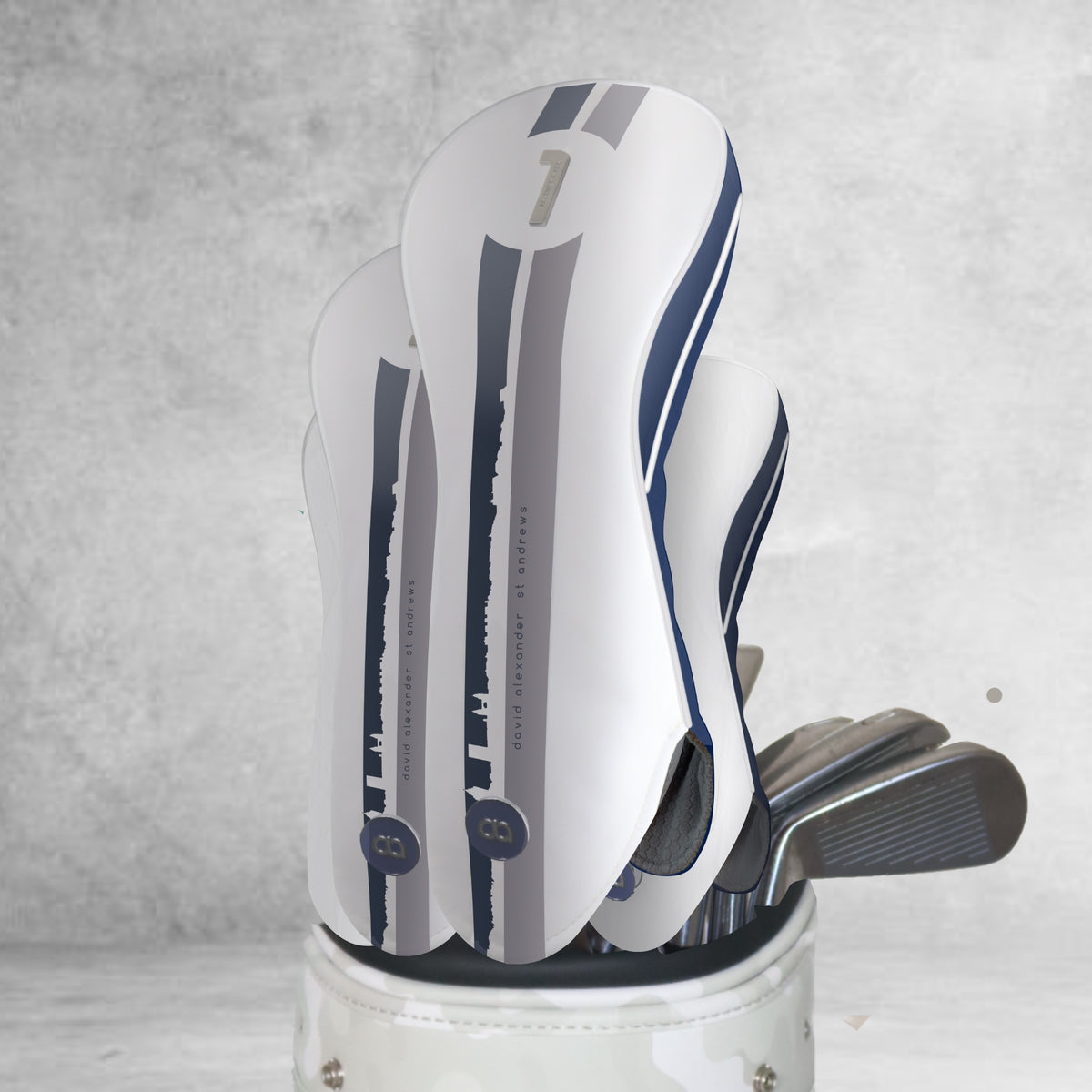A set of white and navy blue leather headcovers with two vertical stripes sporting a St. Andrews cityscape design. Shown on a light grey background