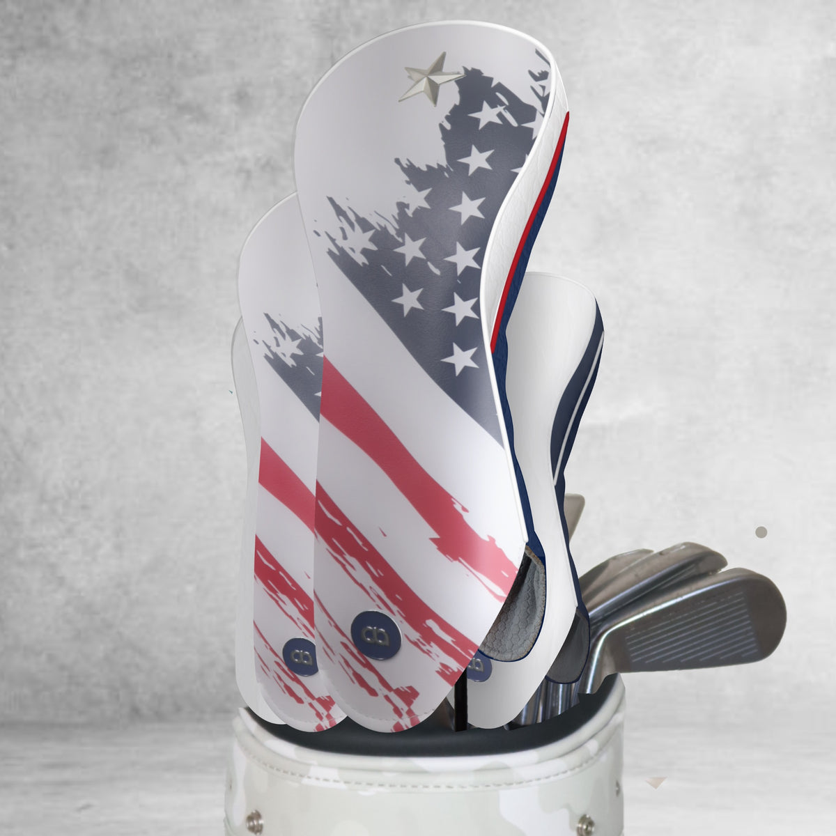 A golf club headcover set in a white golf bag, featuring a grunge American flag design with red and white stripes and a blue field with white stars. The headcovers are white with red and blue accents, and the club heads are visible inside the bag.