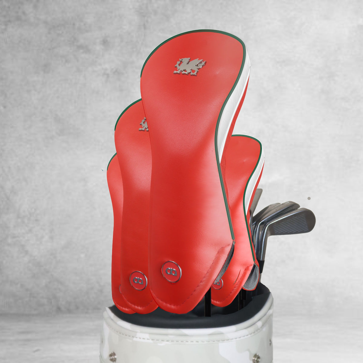 Four red leather Wales golf club headcovers with white and green pipping and a metal dragon logo, standing upright in a white golf bag against a gray concrete wall.