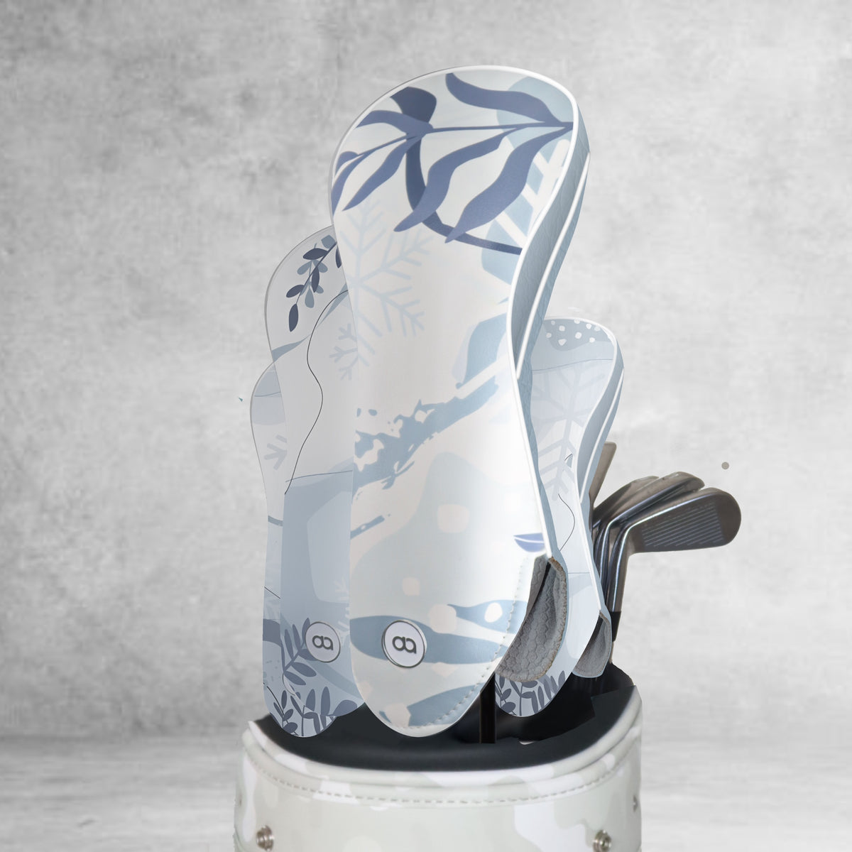 A golf club set in a white and gray patterned golf bag, featuring light blue and white winter-themed leather golf club covers with snowflake and leaf designs, set against a textured gray background.