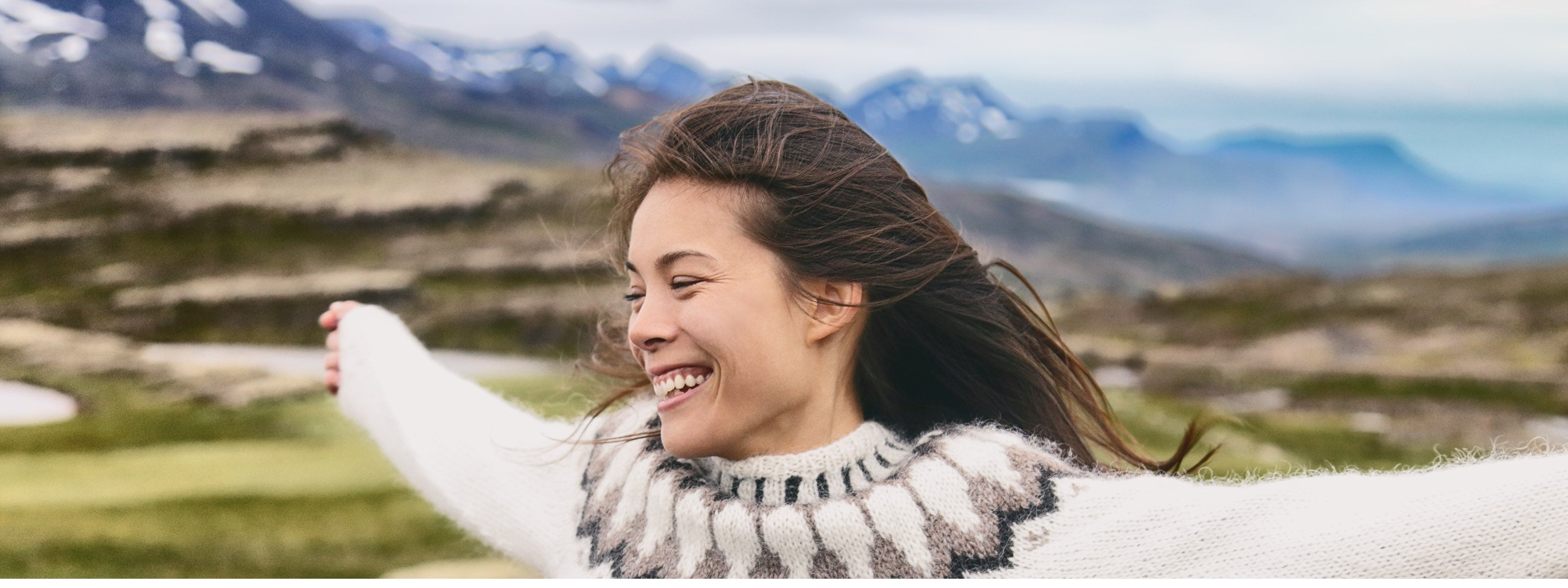 Woman in a striped sweater standing in a scenic outdoor setting with mountains and greenery.