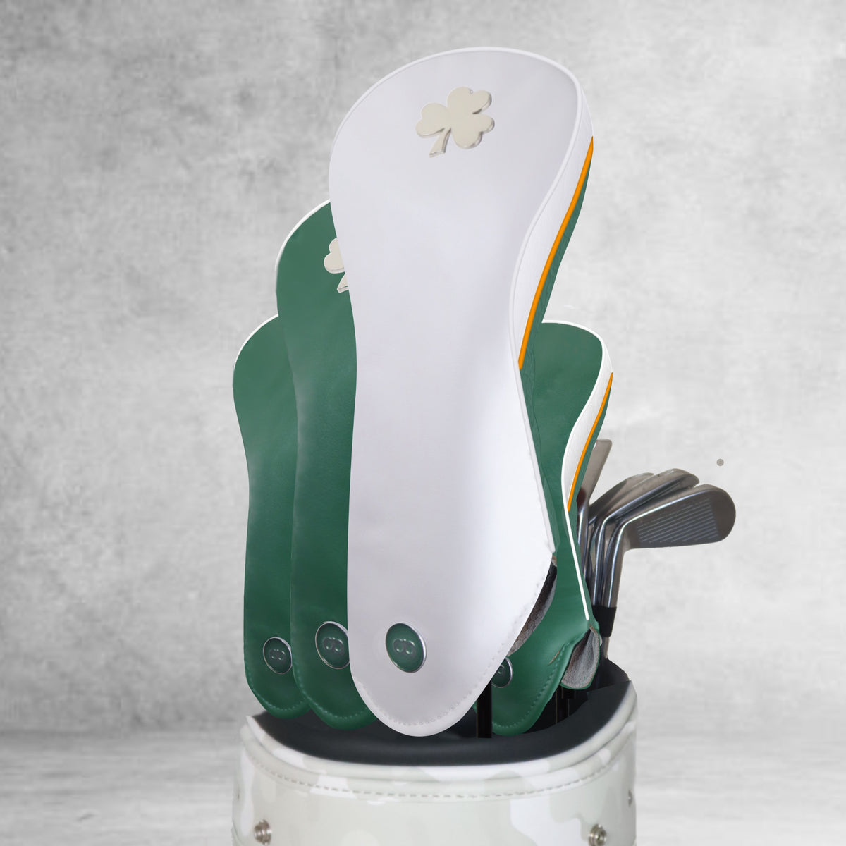 A golf bag filled with three green and white leather Ireland golf headcovers featuring a metal shamrock logo, set against a textured gray background
