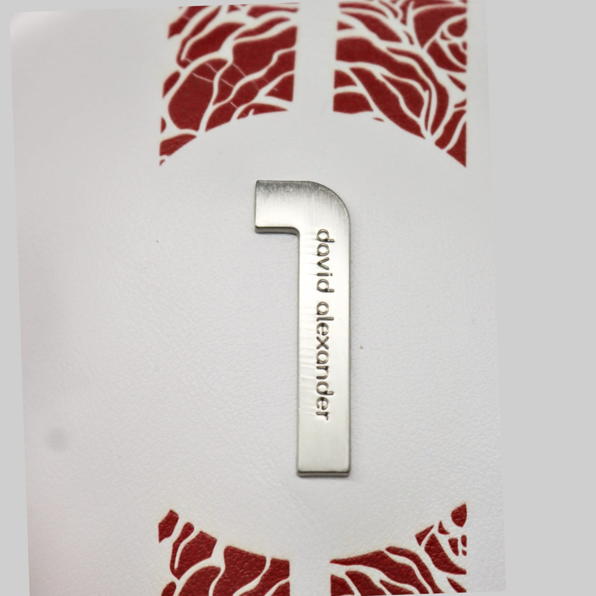 A silver metal Hebrew letter 'Shin' engraved with the name 'David Alexander' vertically, displayed on a white textured card with red decorative patterns.