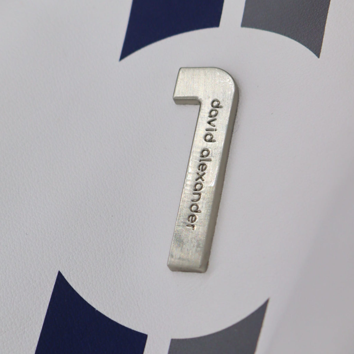 A close-up of a chrome metal  number engraved with the name 'david alexander' on a white background with dark blue and gray graphic shapes.