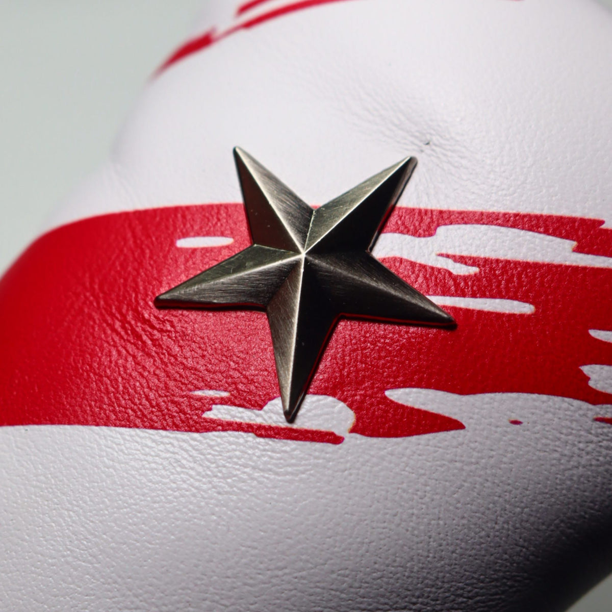 A close-up of a white USA leather golf club putter headcover featuring a bold red horizontal stripe with a distressed, paint-splatter effect and a chrome silver five-point star emblem in the center.