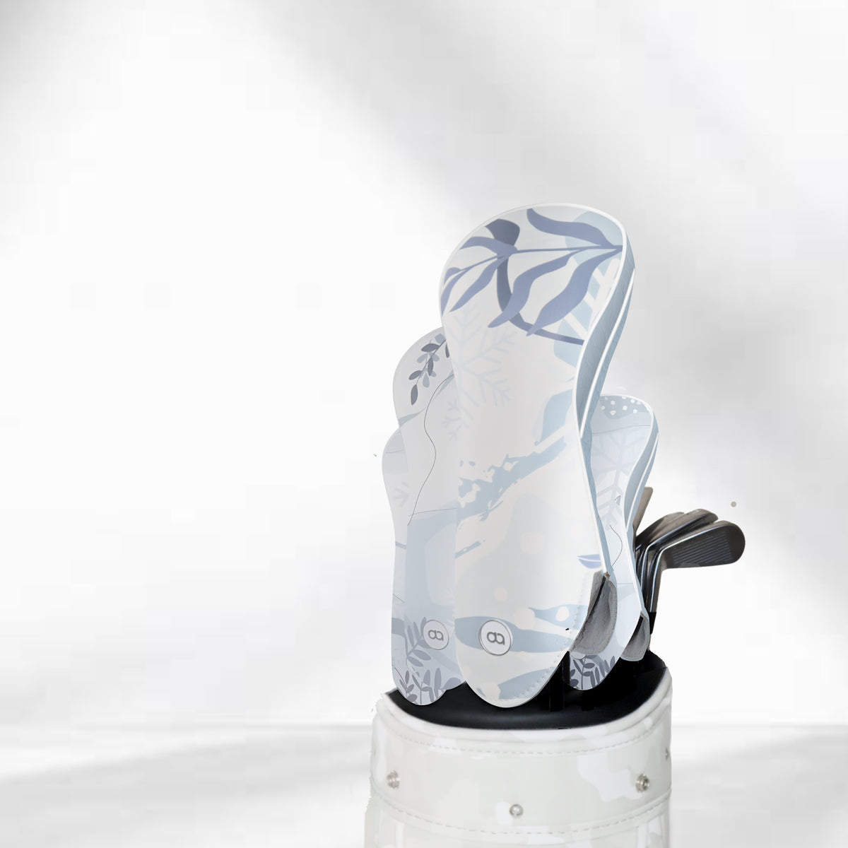 A golf club set in a white and gray patterned golf bag, featuring light blue and white winter-themed leather golf club covers with snowflake and leaf designs, set against a textured gray background.